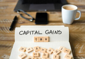 Closeup on notebook over vintage desk surface, front focus on wooden blocks with letters making Capital Gains Tax text. Business concept image with office tools and coffee cup in background